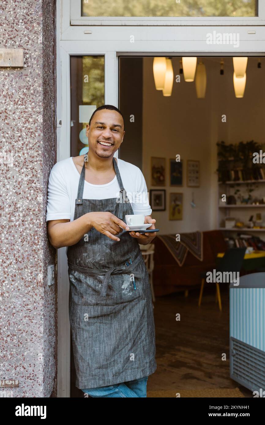 Smiling male owner with coffee cup leaning on wall near cafe doorway Stock Photo