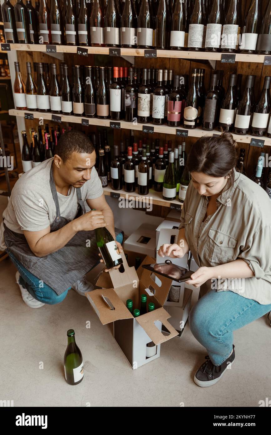 Male and female owners checking inventory of bottles while crouching in wine store Stock Photo
