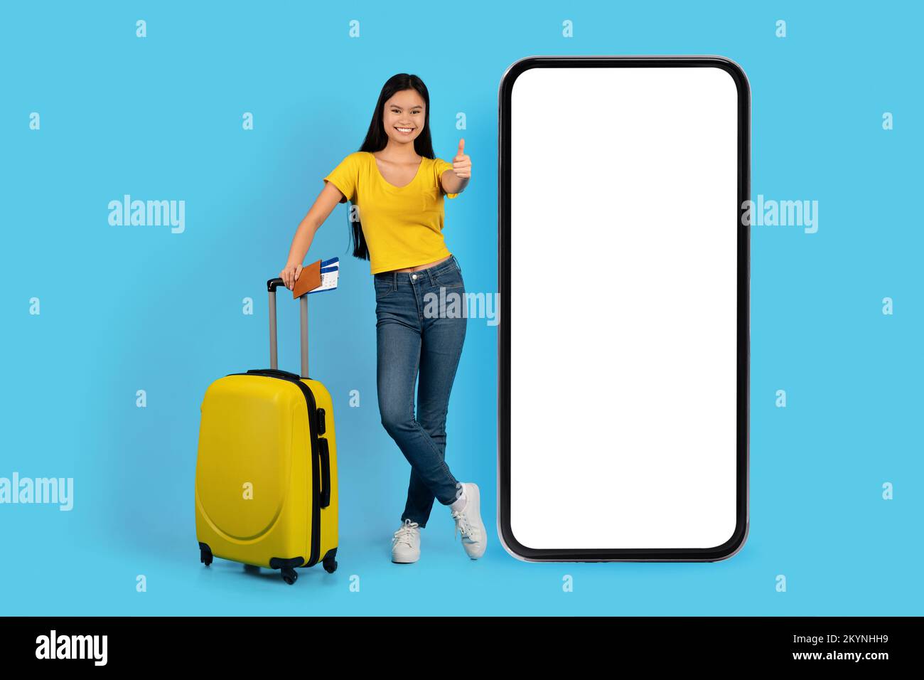 Cheerful young japanese lady in yellow t-shirt with suitcase, tickets show thumb up near huge phone Stock Photo