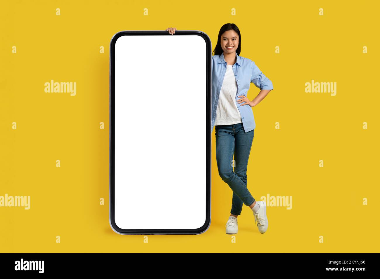 Cheerful millennial korean lady student in casual stand with huge smartphone with blank screen Stock Photo