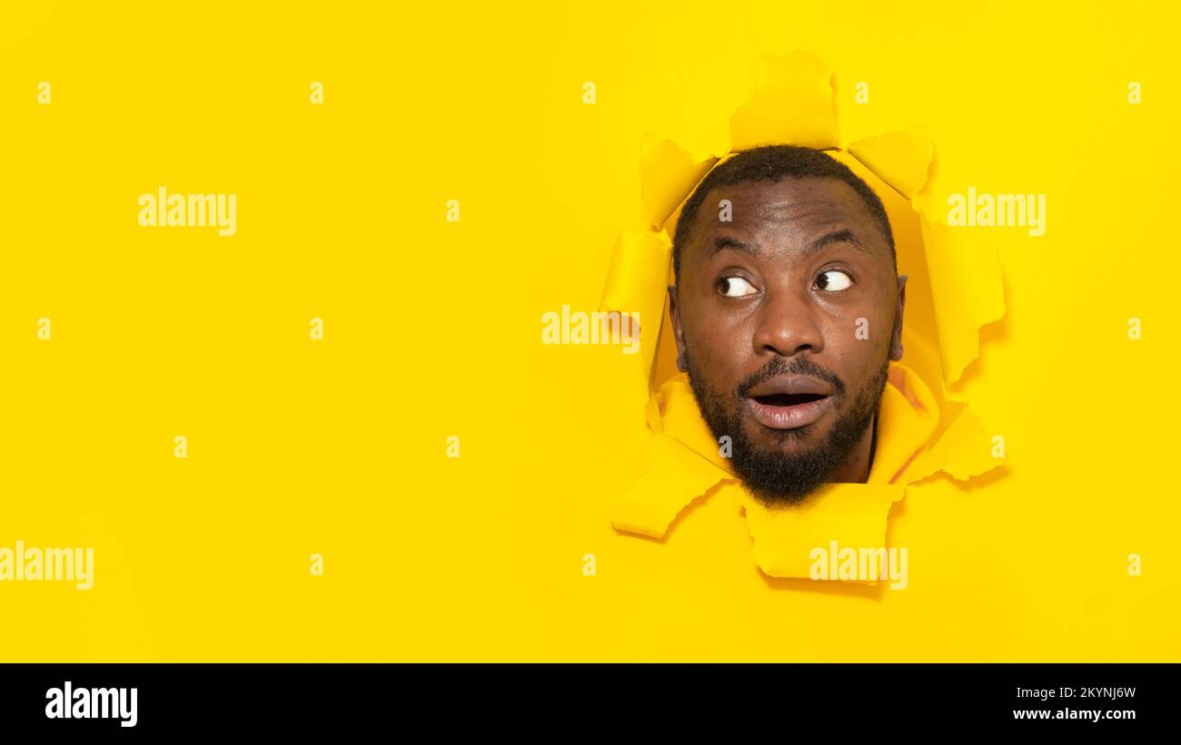 Omg. Shocked black man looking aside at free space through hole in torn yellow paper, posing with opened mouth, panorama Stock Photo
