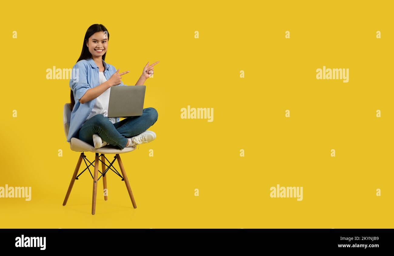 Glad cute millennial korean lady student in casual show fingers at empty space, sit on chair with laptop Stock Photo