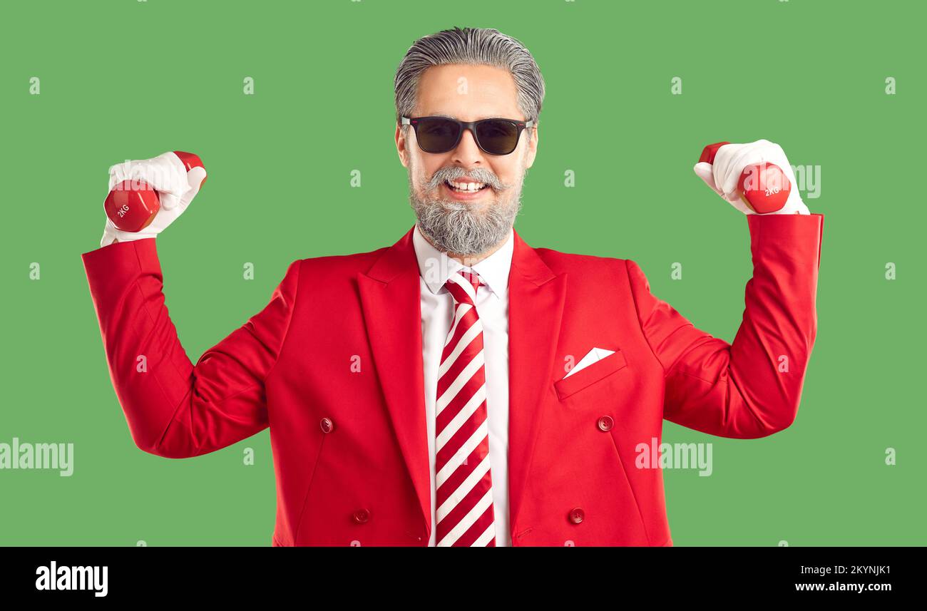 Funny modern Santa Claus in a stylish red suit and striped tie holds dumbbells showing how strong he is. Stock Photo