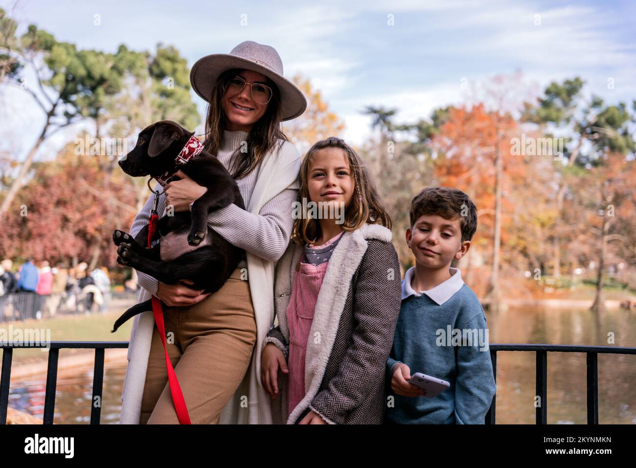 Happy mom in outerwear hugging Labrador Retriever puppy with leash and smiling while standing near children on bridge over pond on weekend day  Stock Photo