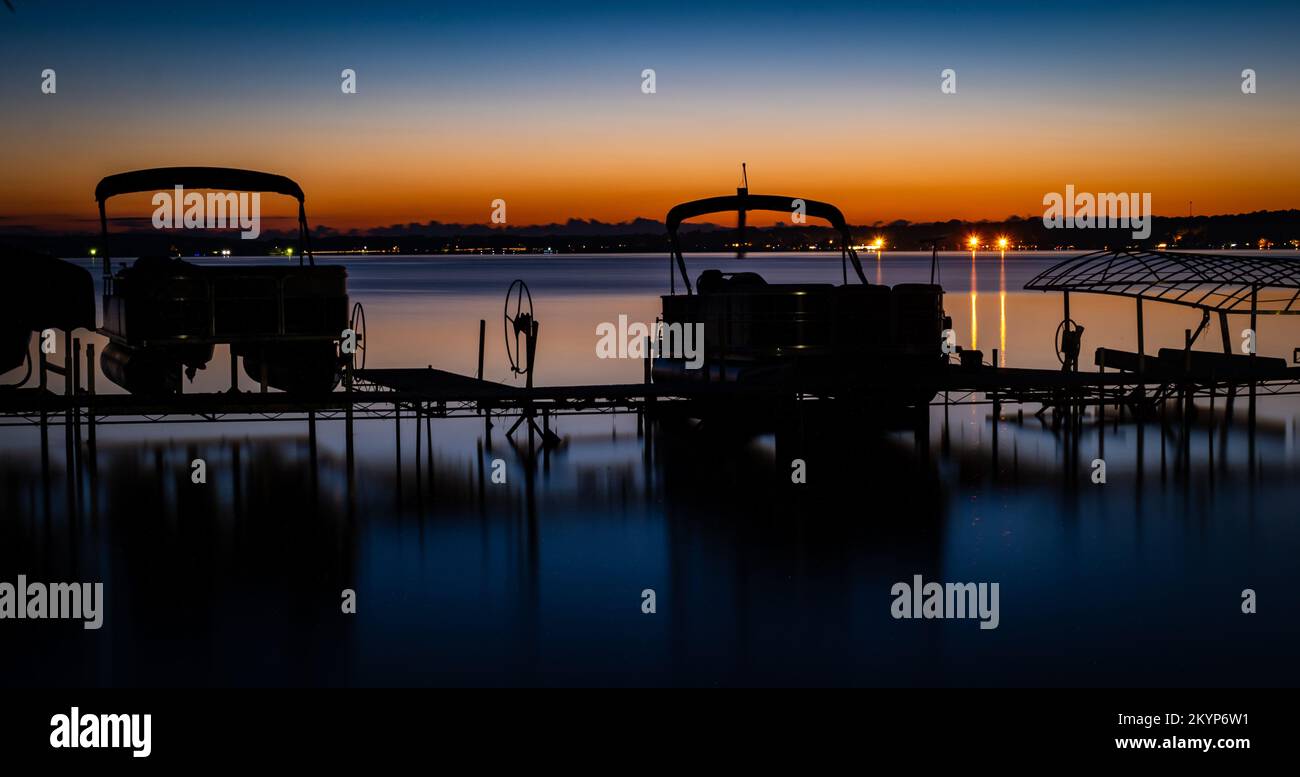 Silhouette of pontoons on boat lifts at lakeshore with an orange sunset in the distance at dusk. Stock Photo