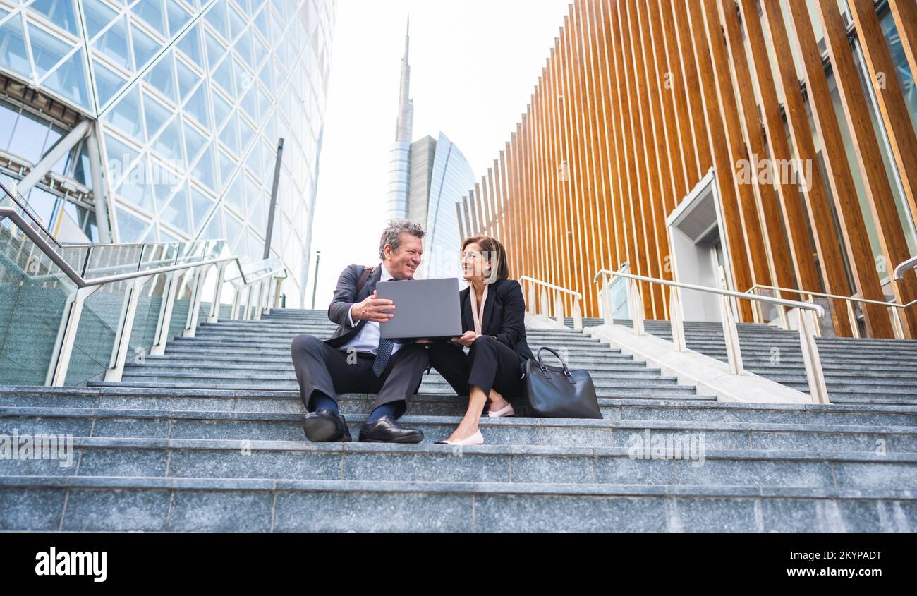 Business couple working on computer while sitting together on the stairs of finance buildings as background - Two business people analyzing financial Stock Photo