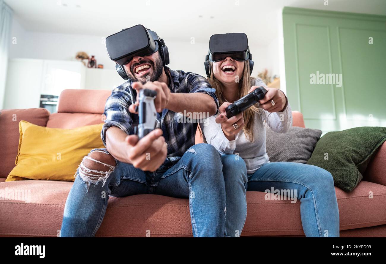 Happy couple playing video games with virtual reality glasses - Portrait of couple in VR glasses playing video game with joystick . Stock Photo