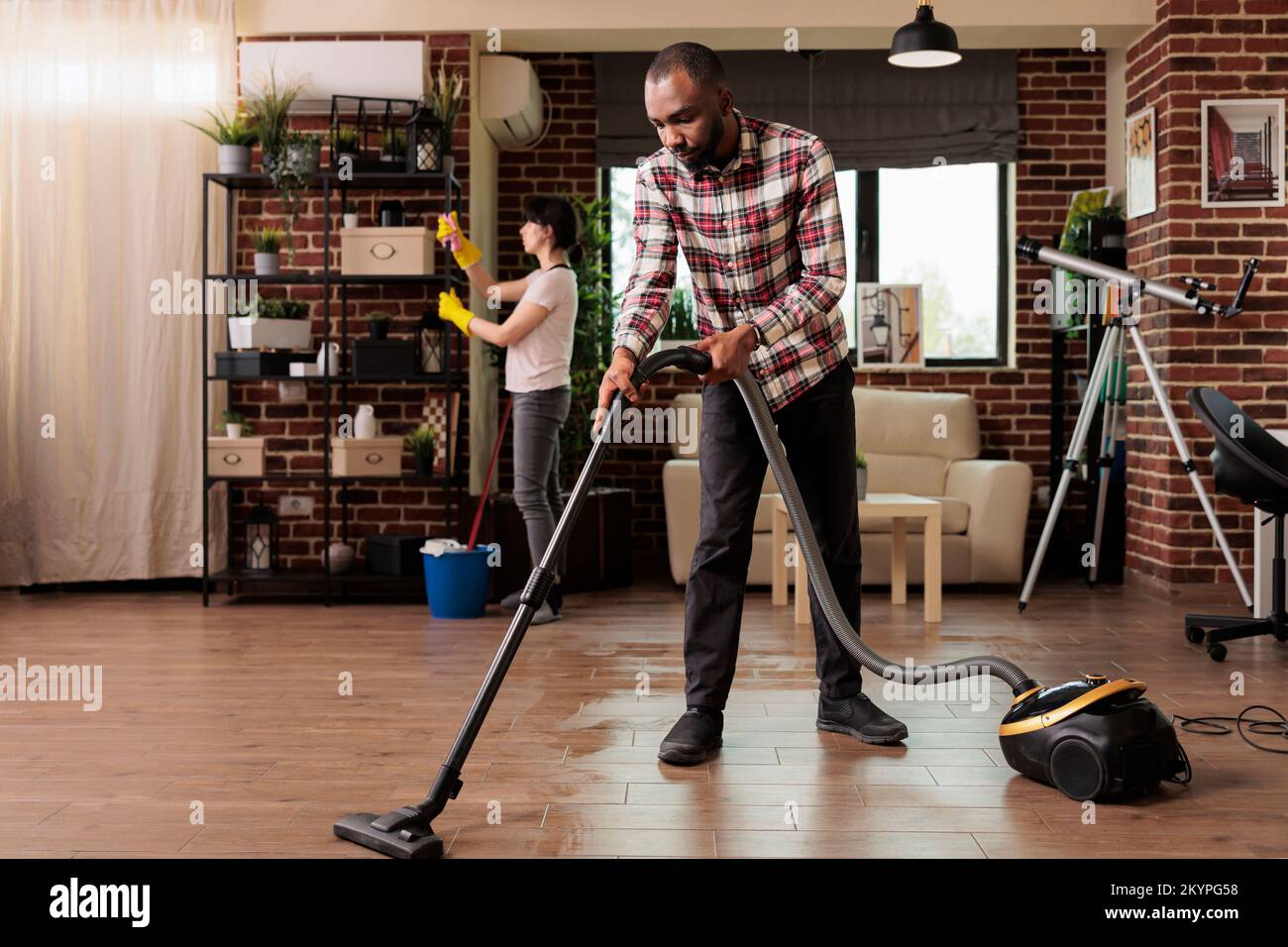 African american man vacuuming floor at home while female roommate dusts shelves in background. Woman wearing rubber gloves while cleaning urban apartment, cleaning day. Stock Photo