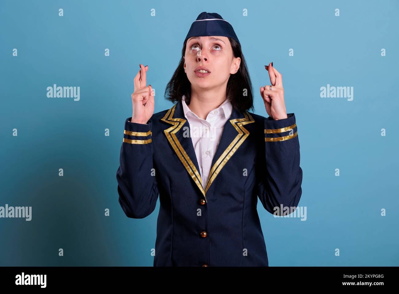 Flight attendant crossing fingers, looking up, praying for luck before flying. Superstitious stewardess in uniform getting lucky, pleading for success with hopeful facial expression Stock Photo