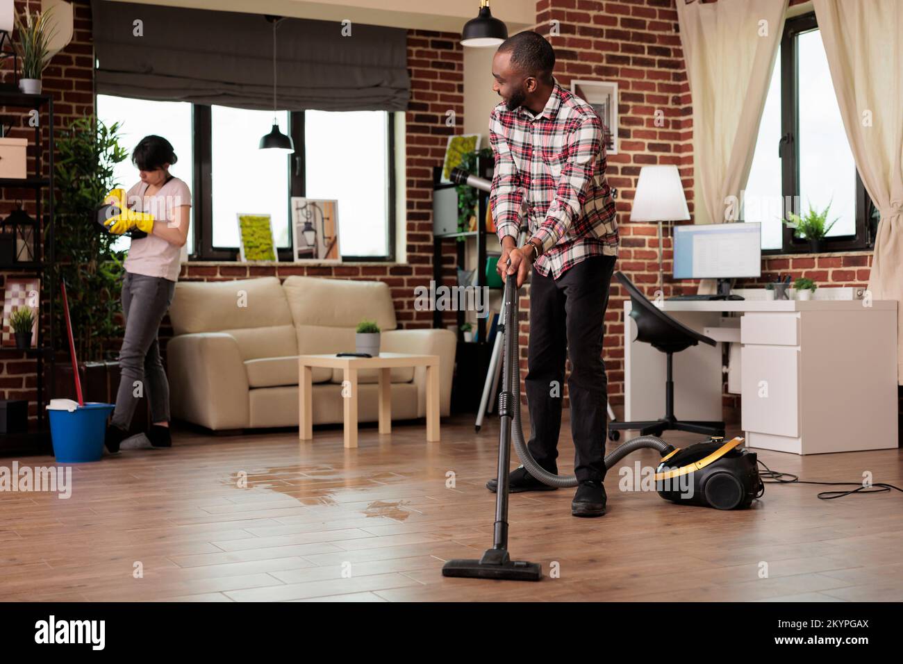 Happy african american man vacuuming floor while concentrated woman dusts decorative objects. Interracial couple cleaning home in harmony, doing household chores together. Stock Photo