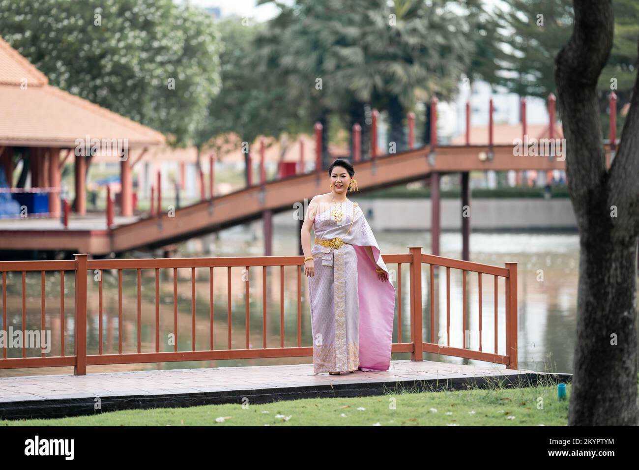 A woman in a traditional Thai costumes is standing on the wooden bridge while being taken a portrait photo shooting. Stock Photo