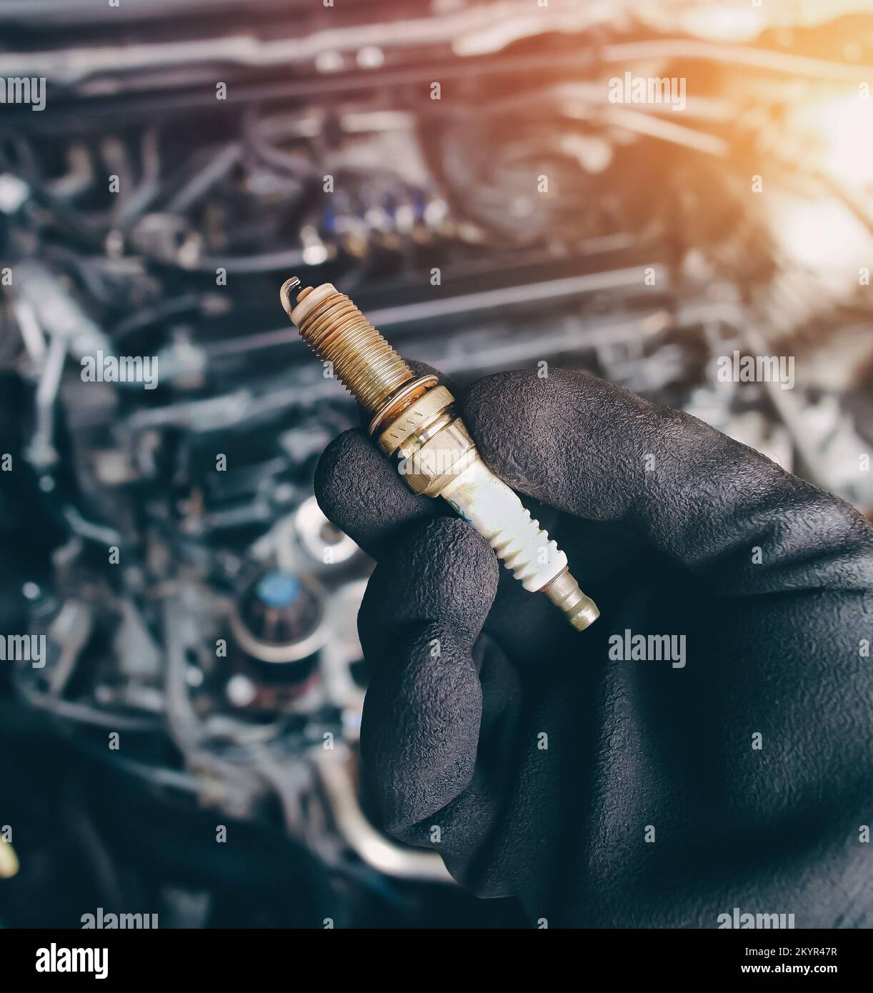 Automobile iridium spark plugs holds by auto mechanic hand with engine compartment blurred background and sunlight. Stock Photo
