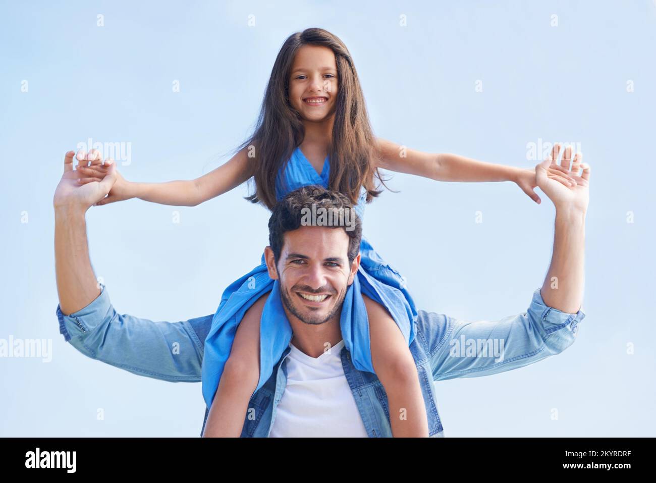 When Im with Daddy, Im on top of the world. Low angle portrait of a girl sitting on her fathers shoulders against a blue sky. Stock Photo