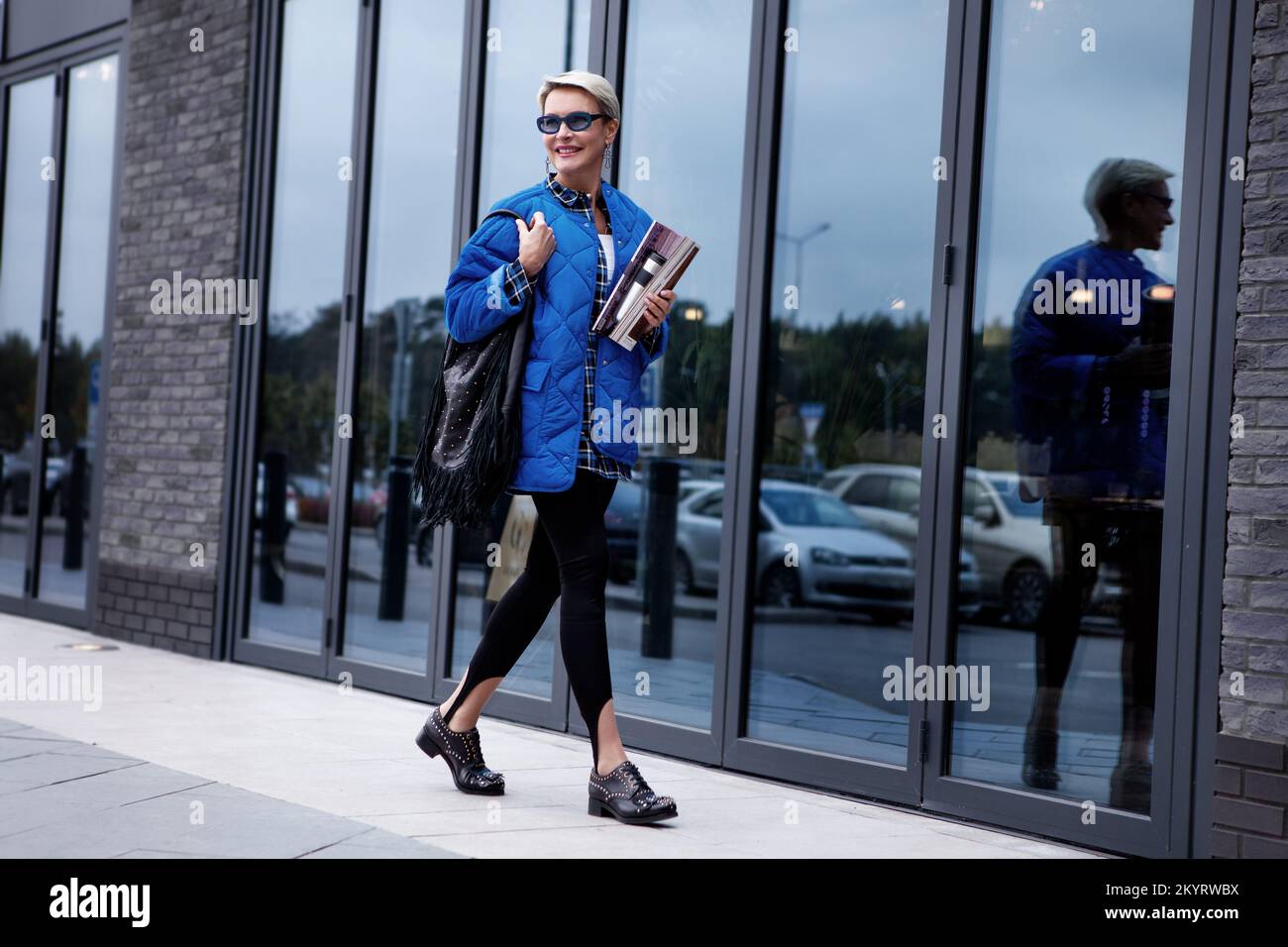 Fashionable woman with short hair walking street dressed in stylish casual outfit, blue colorful jacket and sunglasses, black leather handbag and boot Stock Photo