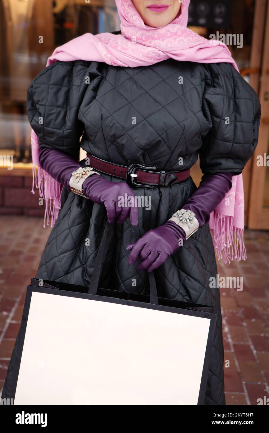 Woman holding shopping bag, wearing stylish leather gloves and bracelets, elegant head scarf and quilted coat. Fashionable female clothing and accesso Stock Photo