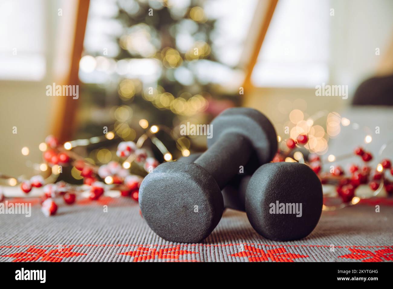 Healthy lifestyle during Christmas feasting period concept. Black dumbbells with Christmas ornaments blurred Christmas tree with Christmas lights on. Stock Photo