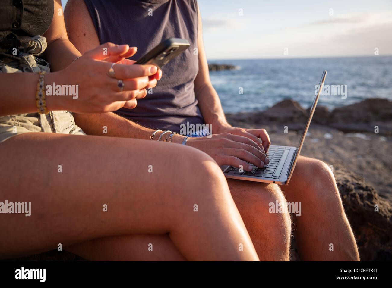 Close-up of two people's hands at the beach using their smartphone and laptop Stock Photo