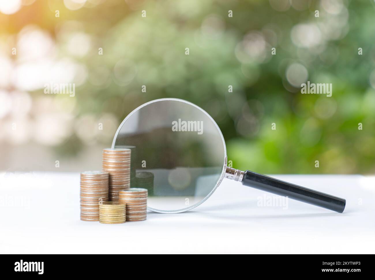 Magnifying glass on the stack of coins on out of focus green tree