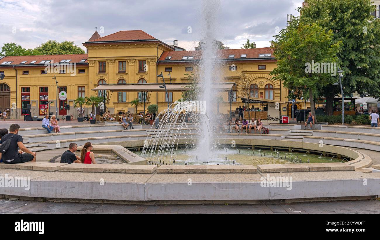 Szeged, Hungary - July 30, 2022: People Sitting Around Water Fountain at Dugonics Square in Front of University Building Summer Day. Stock Photo