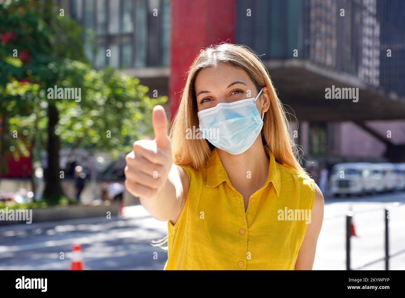 Optimistic young woman wearing protective surgical mask showing thumbs up in Sao Paulo city street, Brazil Stock Photo