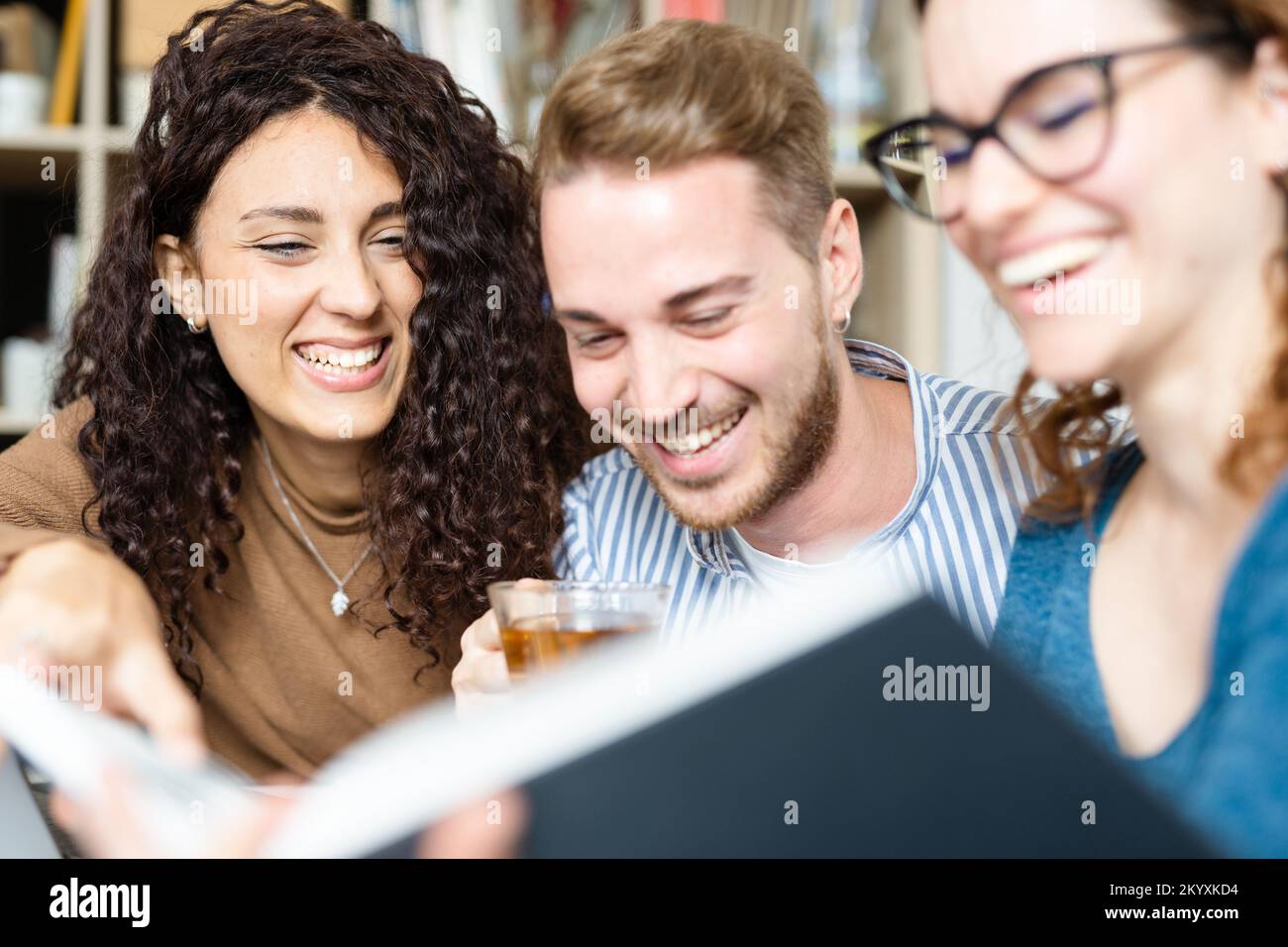 Happy caucasian friends having fun in university library, sitting at table with books while drinking coffee and tea. Stock Photo