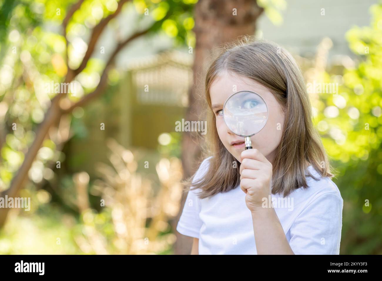 Curious clever elementary school age child, girl looking at the camera through a magnifying glass, holding a loupe in hand, one eye closed, one visibl Stock Photo