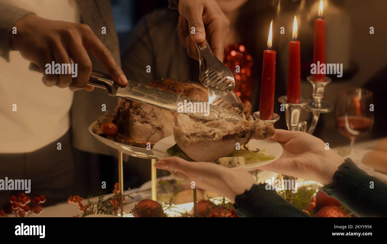 Close-up shot of African American man cutting turkey or chicken. Multi cultural family celebrating Christmas or Thanksgiving Day. Served table with dishes and candles. Family Christmas dinner at home. Stock Photo