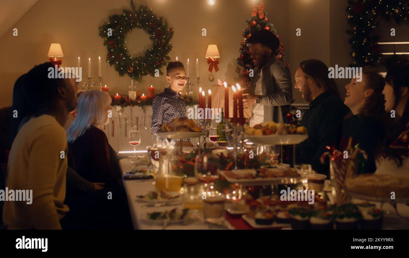 African American man givs gift to young boy. Happy family celebrating Christmas or New Year 2023. Served holiday table with dishes and candles. Warm atmosphere of family Christmas dinner at home. Stock Photo