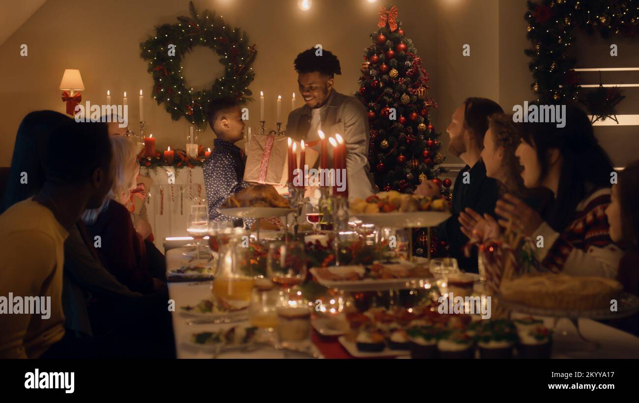 African American man givs gift to young boy. Happy family celebrating Christmas or New Year 2023. Served holiday table with dishes and candles. Warm atmosphere of family Christmas dinner at home. Stock Photo