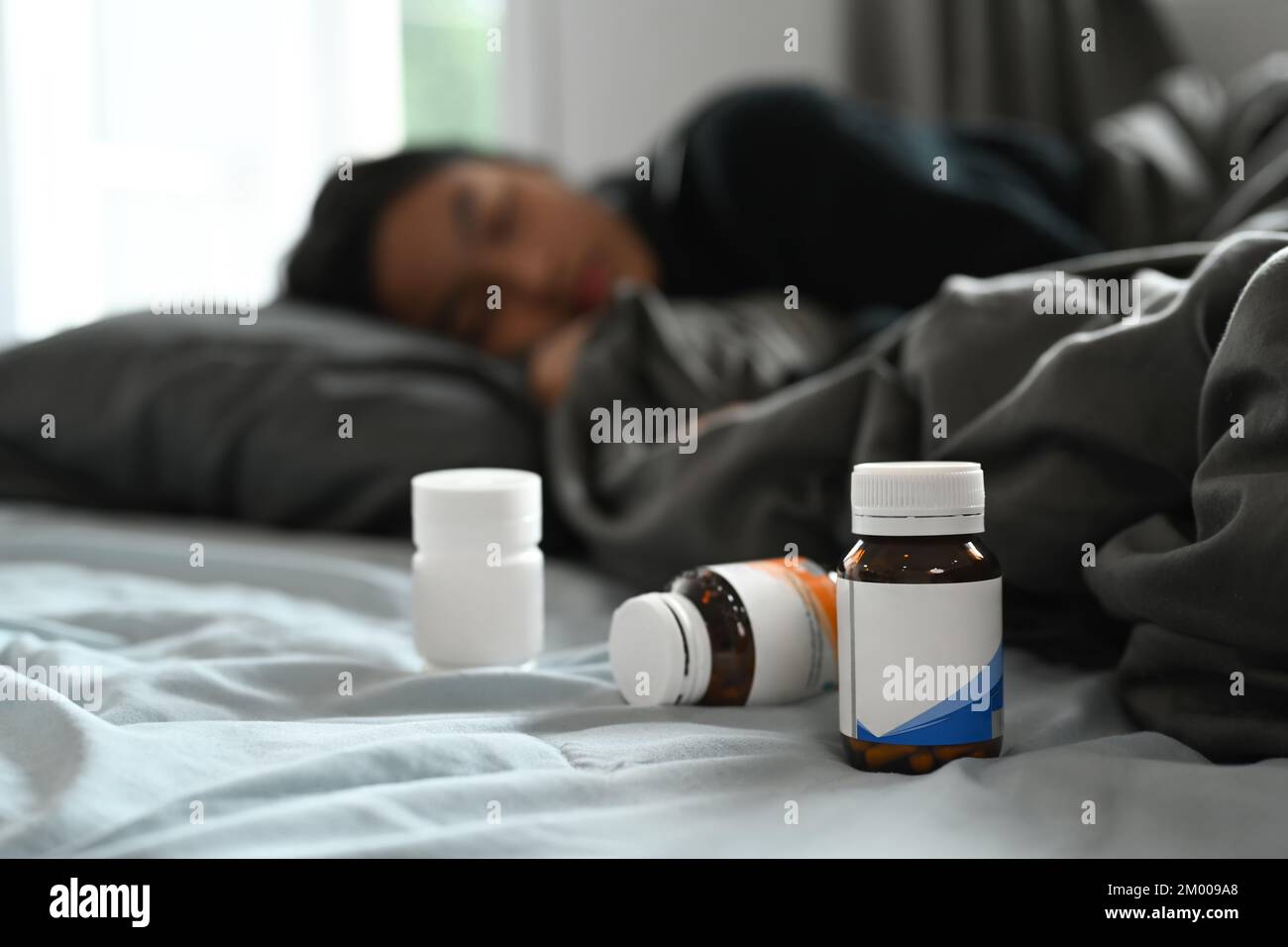 Pill bottles on bed with ill young woman lying under blanket with high fever. Medicine
