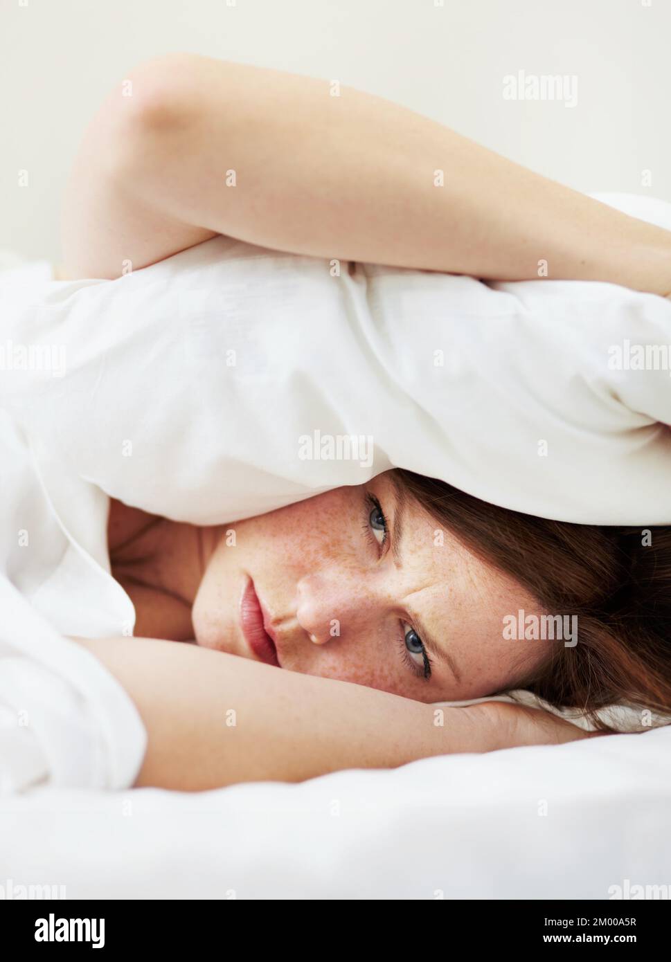 Its too early to be awake. A grumpy looking woman lying in bed with a pillow over her head. Stock Photo