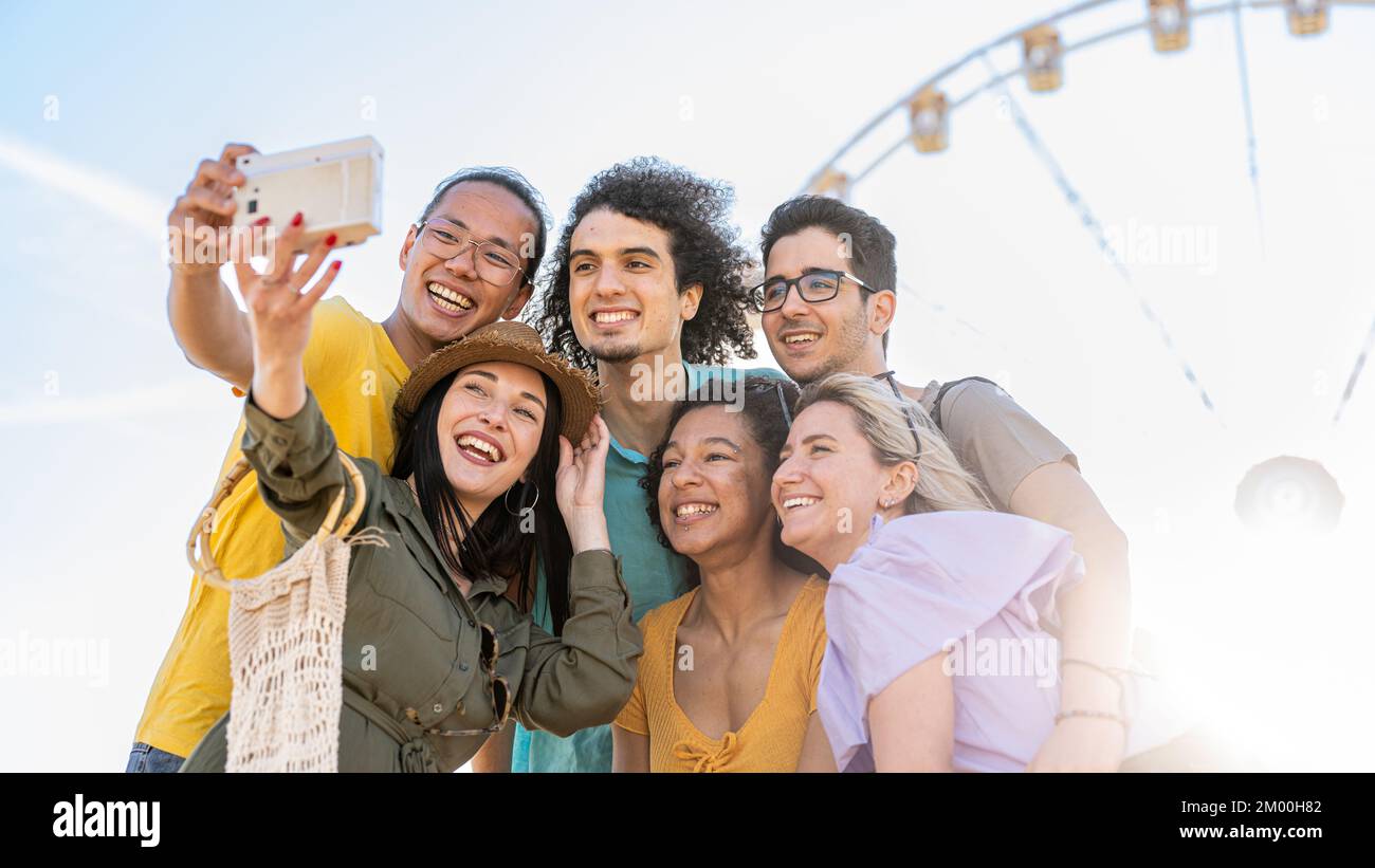 Group of six people having fun taking photos with vintage camera, influencers of generation z creating contents for social media, ferris wheel on back Stock Photo