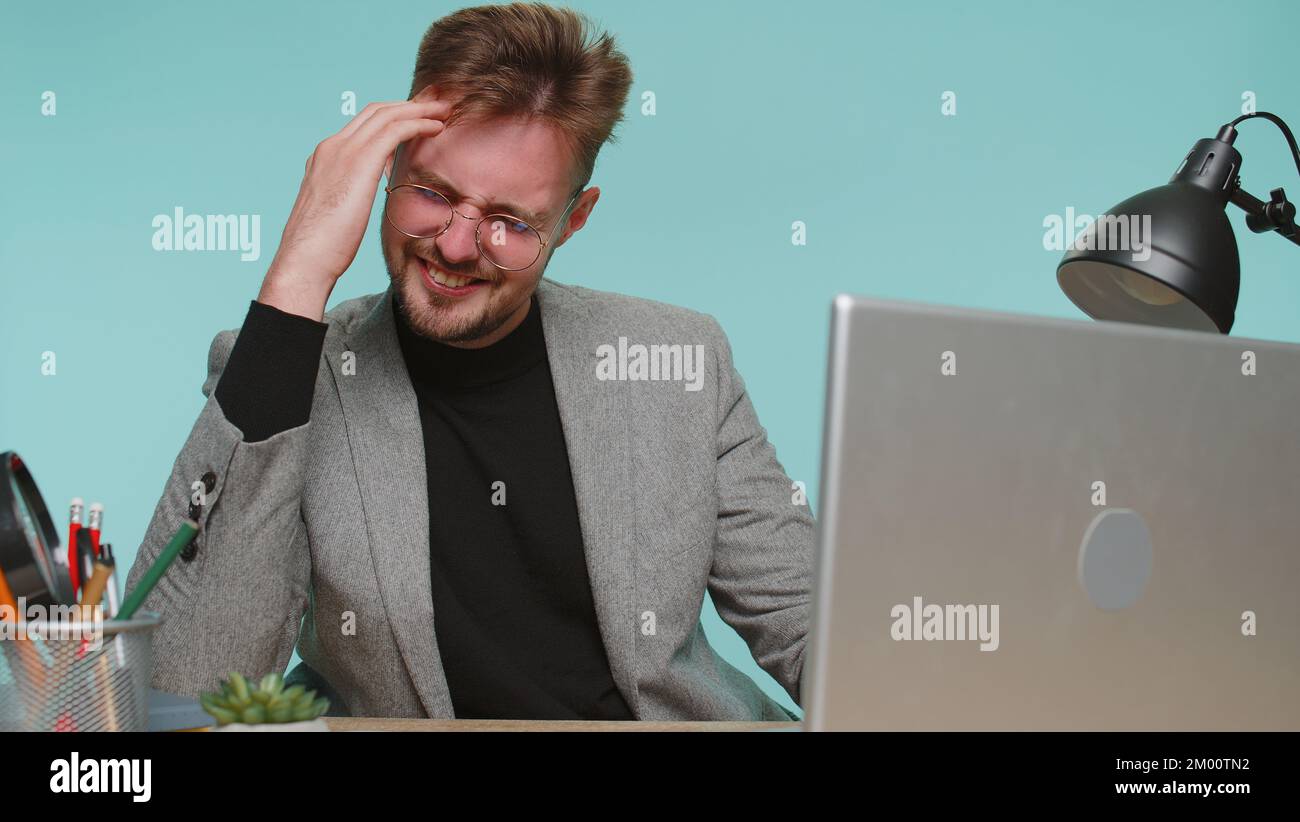 Displeased business man sits at office desk rubbing temples to cure headache problem, suffering from tension and migraine, stress, grimacing in pain, high blood pressure. Young guy on blue background Stock Photo