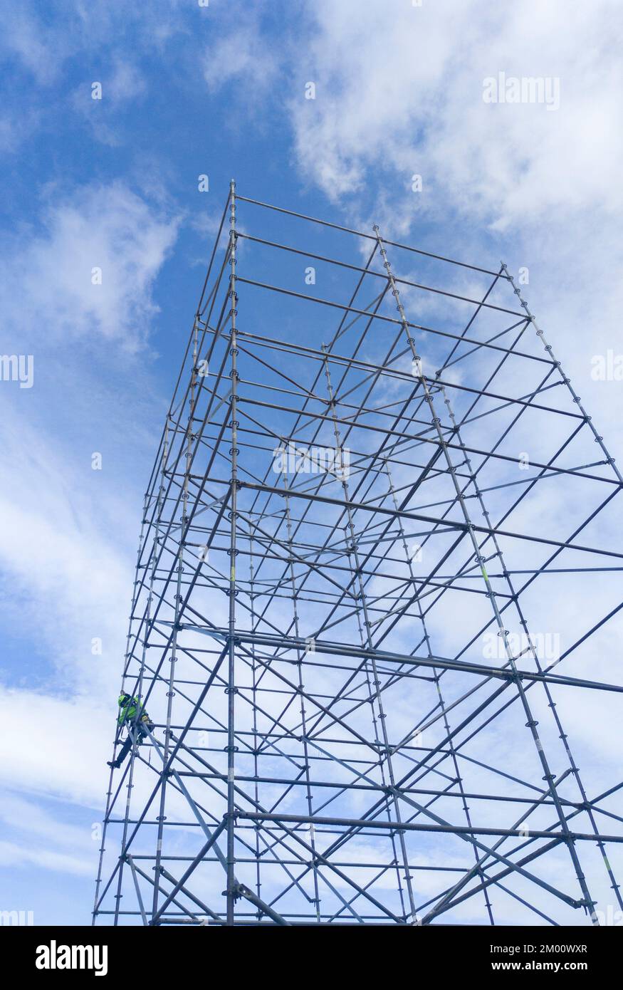 Unidentified worker at height dismantling a scaffolding tower. Blue cloudy sky on background. Stock Photo