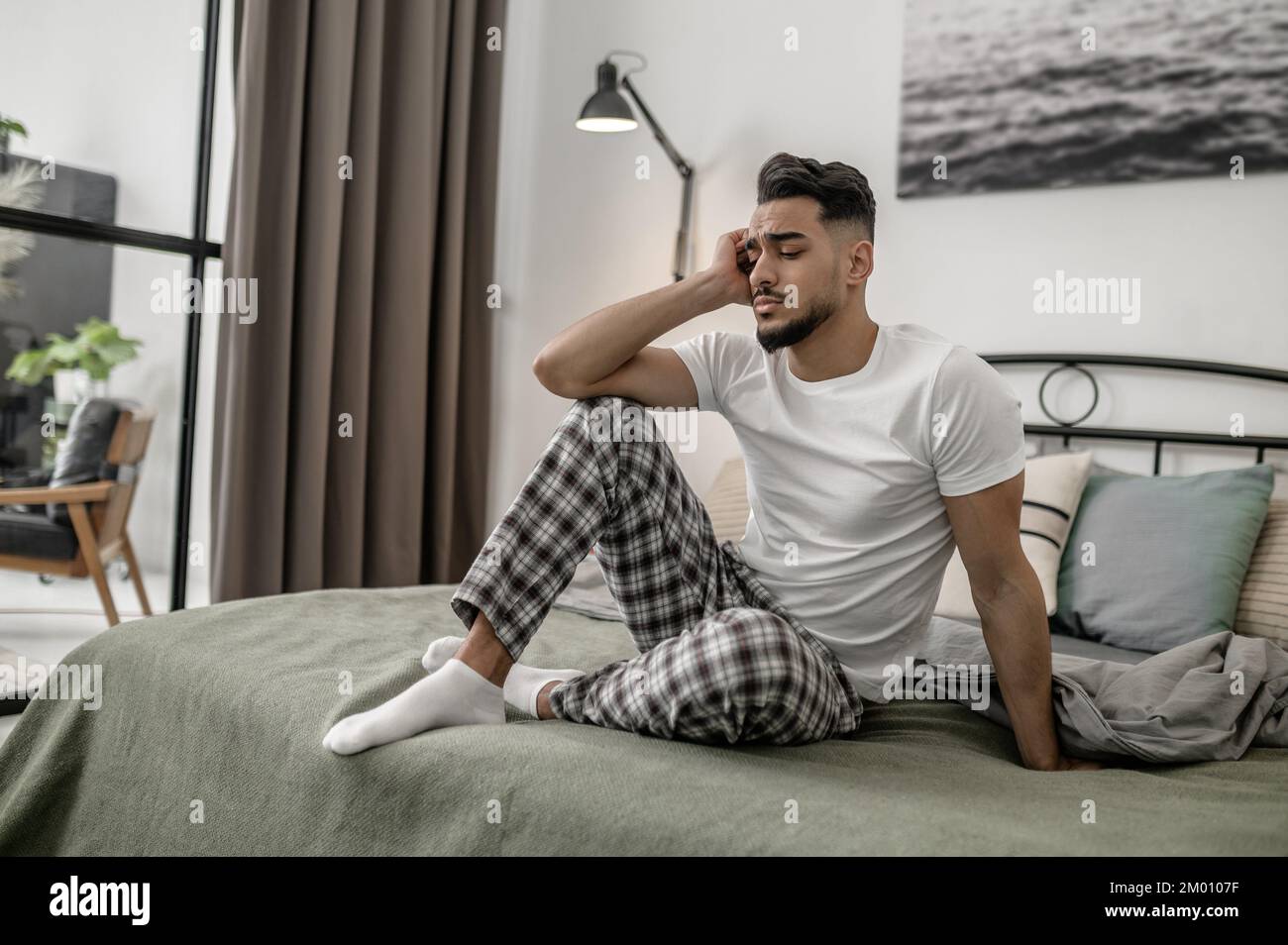 Day off. A man in white tshirt and plaid pants sitting on bed at home. Stock Photo