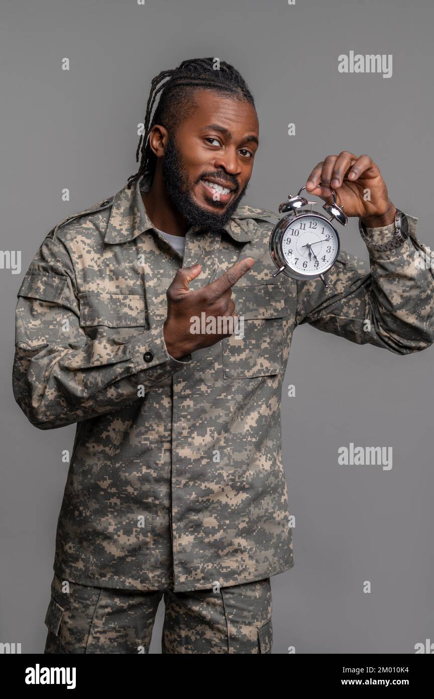Smiling serviceman pointing his index finger at the mechanical wind-up alarm clock with double bells. Stock Photo