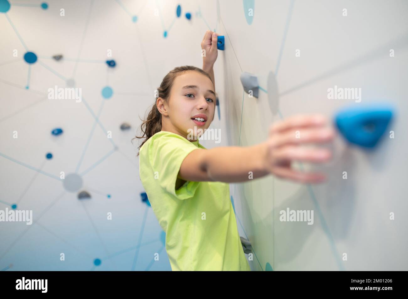 Fortitude. Purposeful striving girl on climbing practice wall with effort stretching her hand towards camera towards ledge in lighted room. Stock Photo
