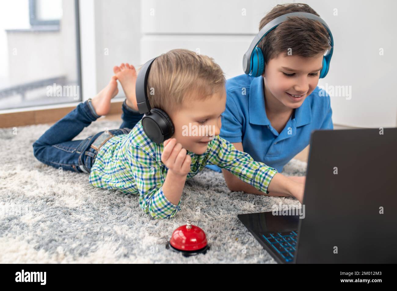 Calm cute little boy and his joyous brother lying on the carpet at their laptop. Stock Photo