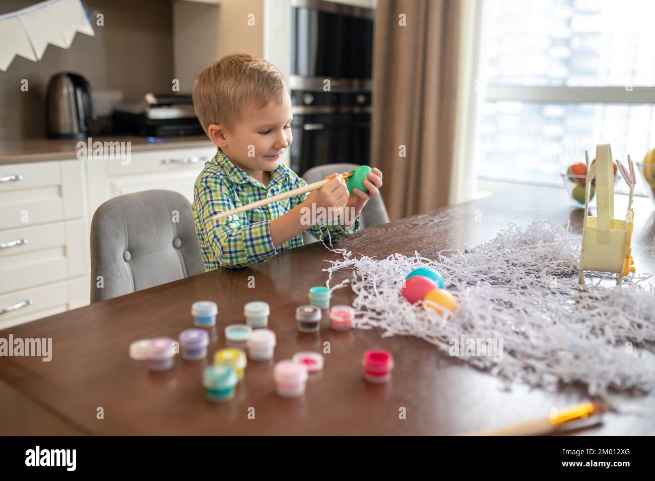 Smiling focused little boy painting a chicken egg with a wooden brush and food coloring. Stock Photo