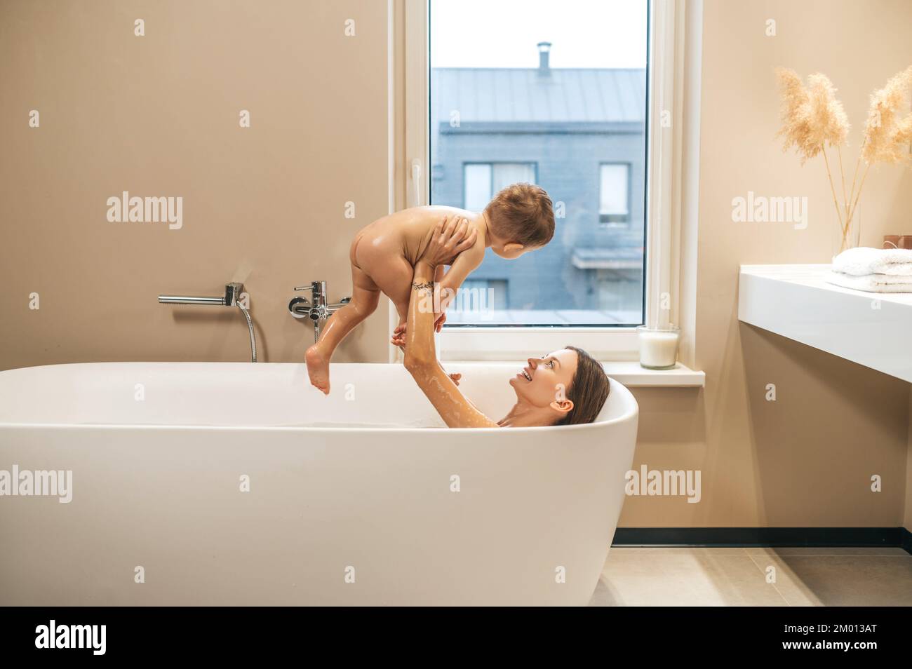 Smiling pleased young woman lying in the bathtub and lifting the baby in the air. Stock Photo