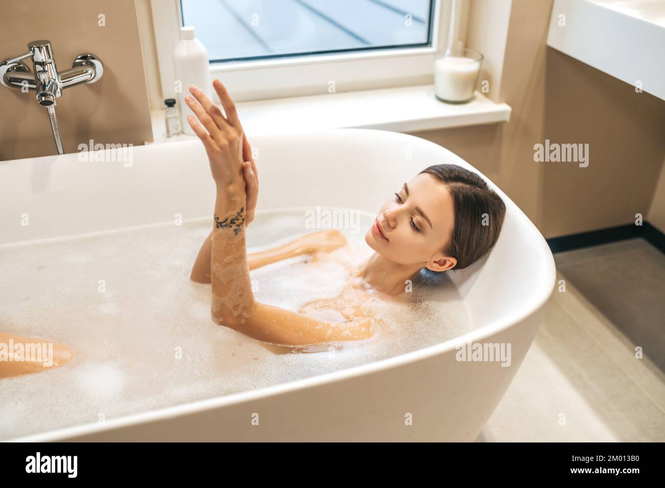Calm lovely young woman lying in the bath and spreading soapy foam over her body. Stock Photo
