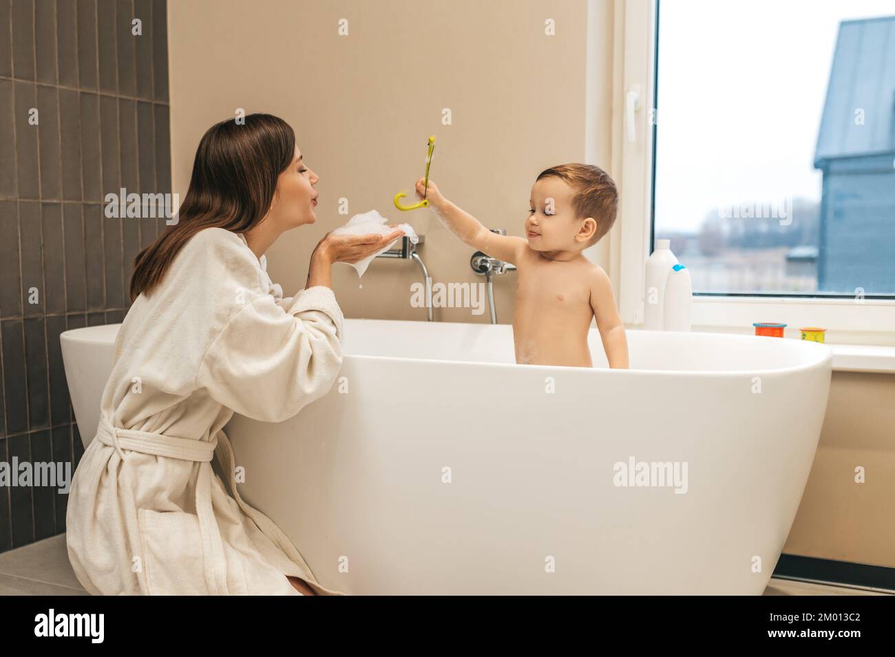 Woman in the bathrobe blowing soapy foam from her palms on her cute child in the bath. Stock Photo