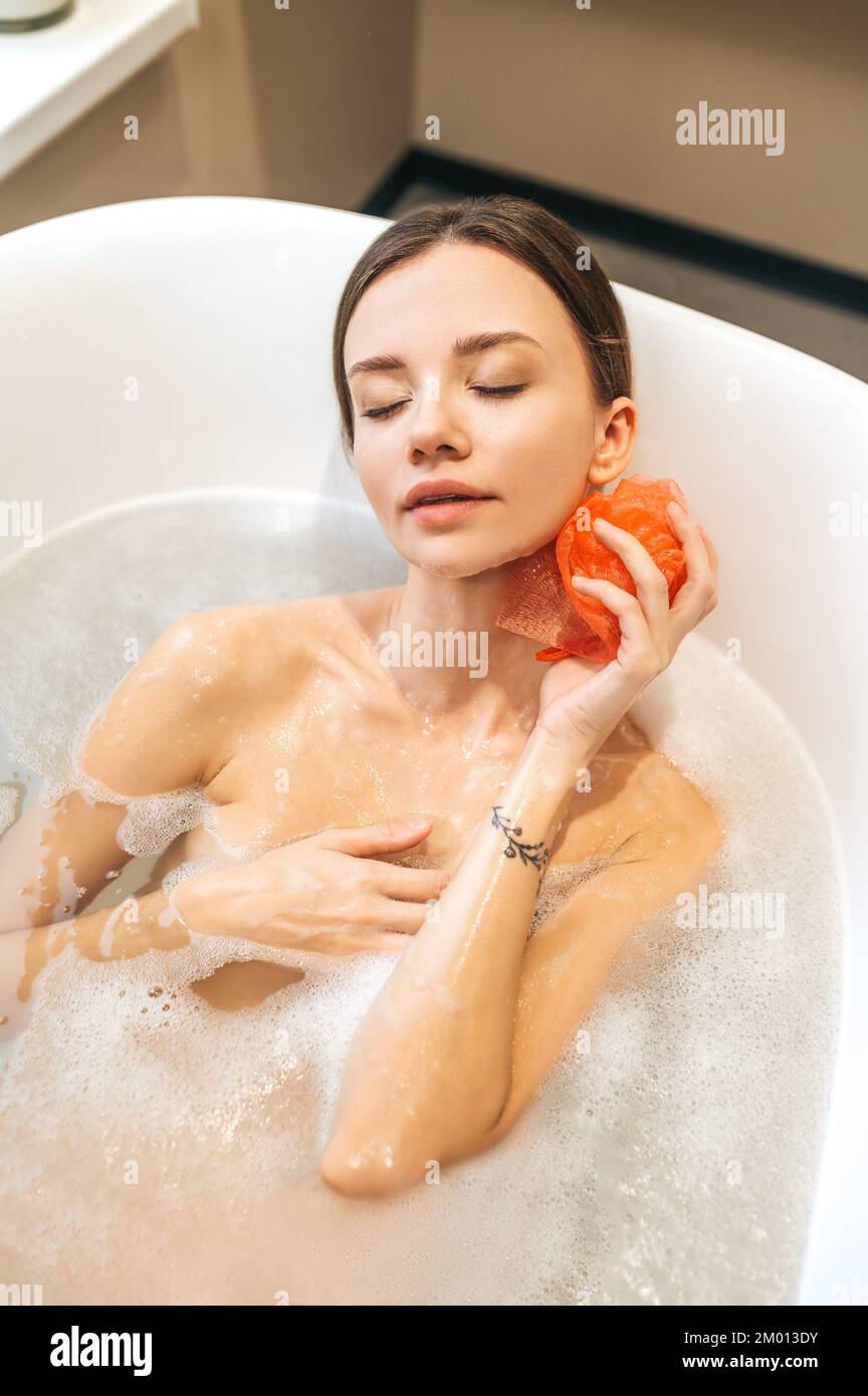 Relaxed lady lying with her eyes shut while washing her body with a soapy nylon loofah. Stock Photo