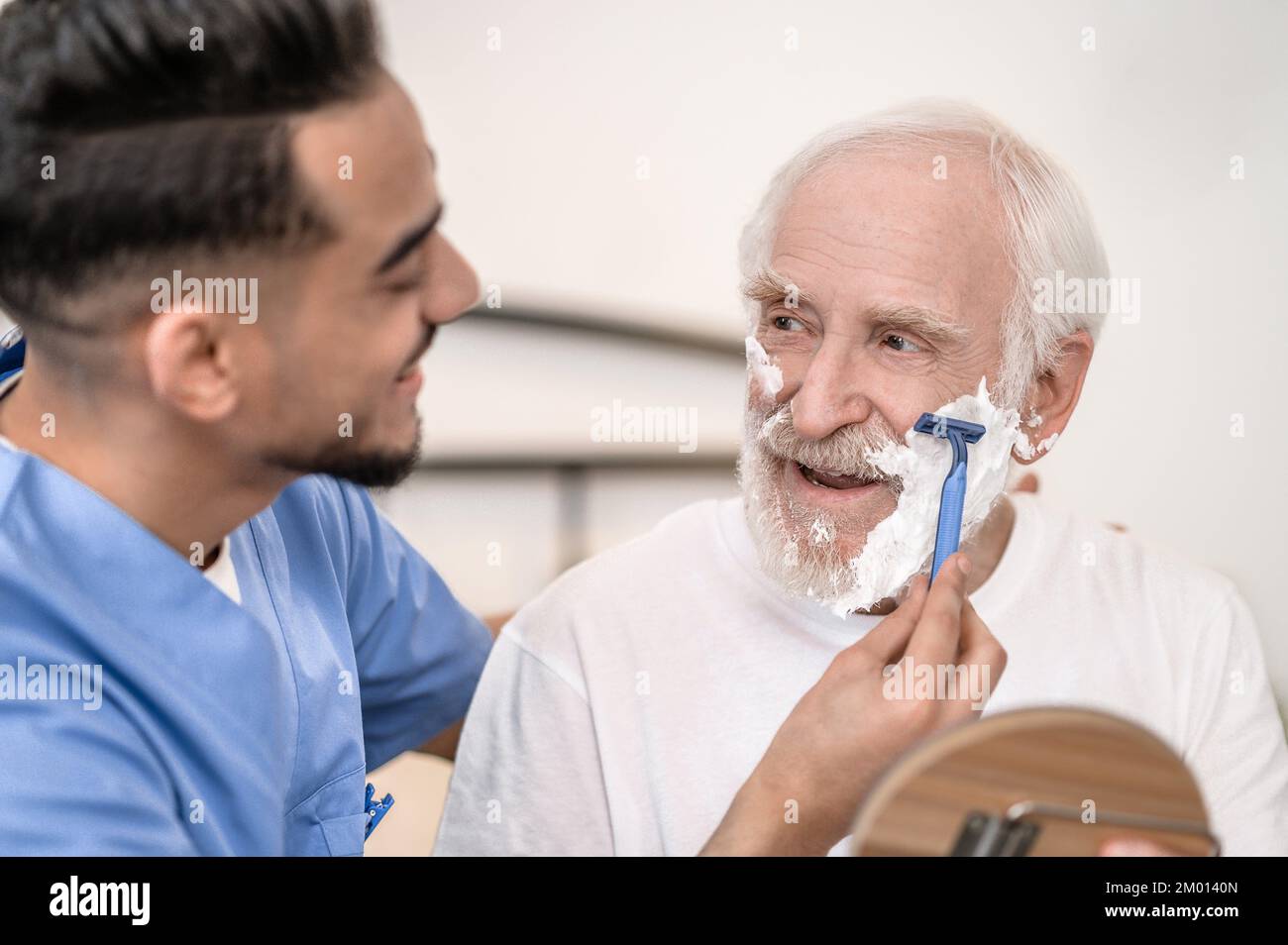 Young caretaker shaving off the stubble on the face of an aged man using a safety razor. Stock Photo