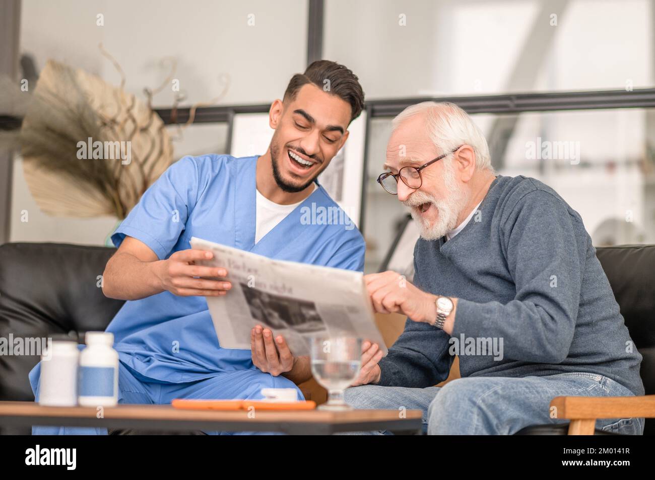 Pleased pensioner and his joyous caregiver sitting at the coffee table and reading the newspaper. Stock Photo