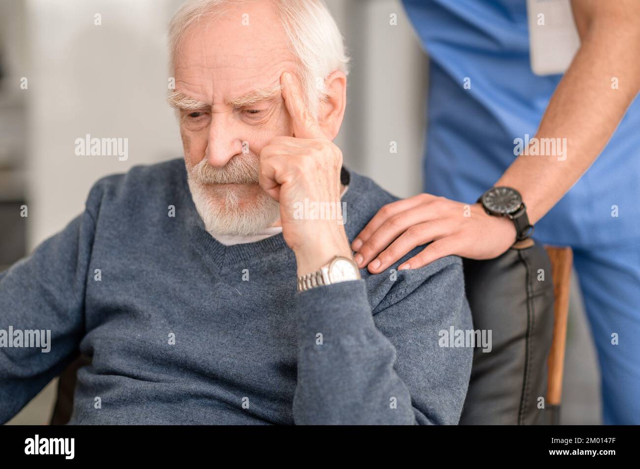 Sad old man sitting in the armchair while his caretaker patting him on the shoulder. Stock Photo