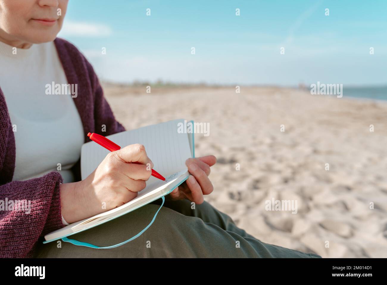Cropped photo of a diarist sitting on the beach by the sea taking notes in her notebook. Stock Photo