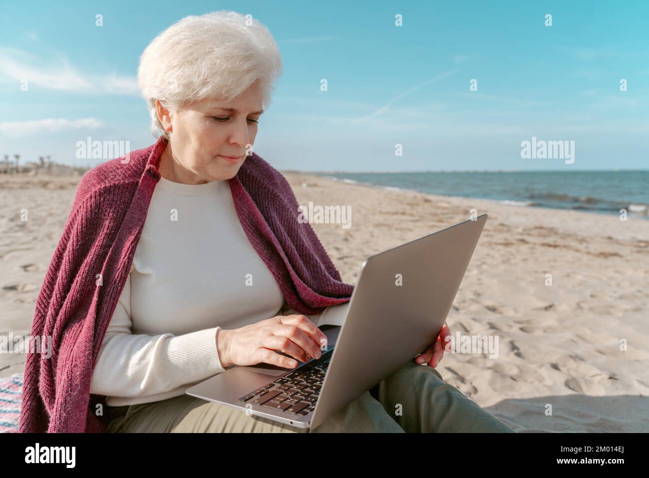 Serious concentrated attractive Caucasian gray-haired lady sitting on the beach and typing on the laptop. Stock Photo