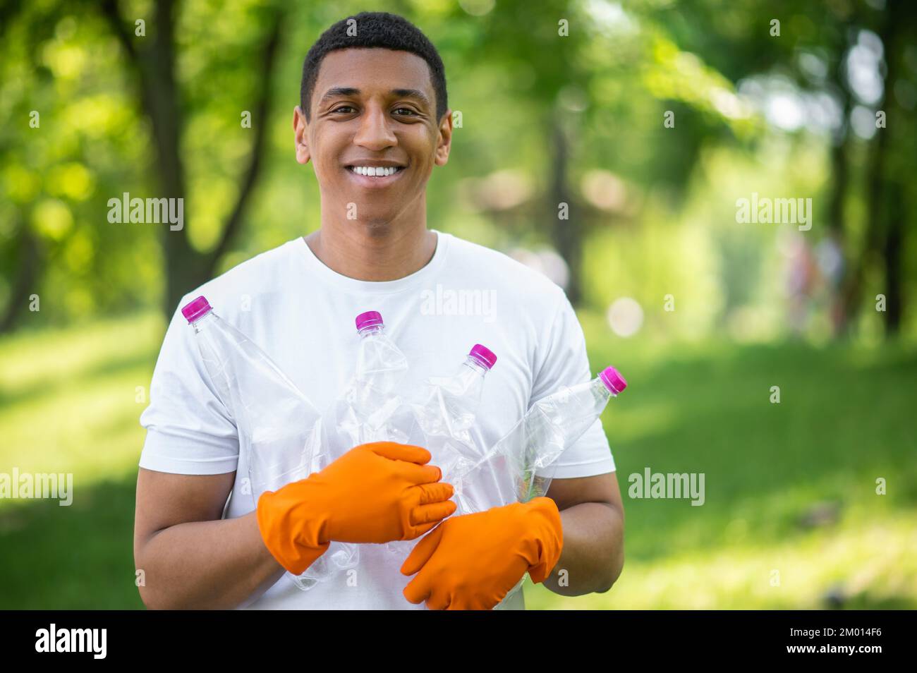 Protection of nature. Dark-skinned guy in gloves smiling confidently at camera holding plastic bottles standing in park on sunny day. Stock Photo