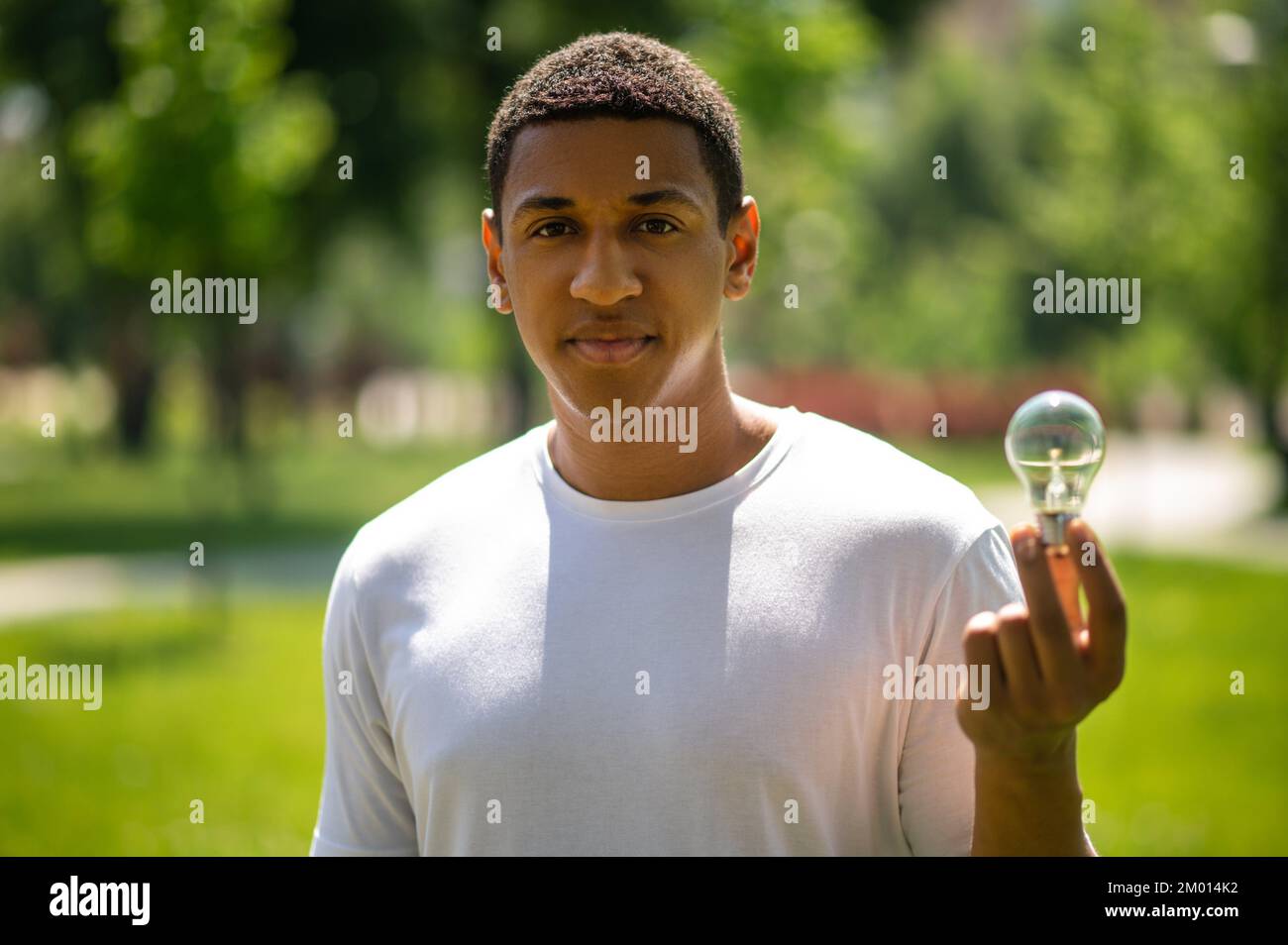 Protection of nature. Dark-skinned guy looking confidently at camera holding lightbulb standing in park on sunny day. Stock Photo